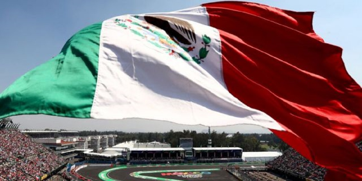 Huge Mexican flag waving over a race track stadium with cheering crowds below.