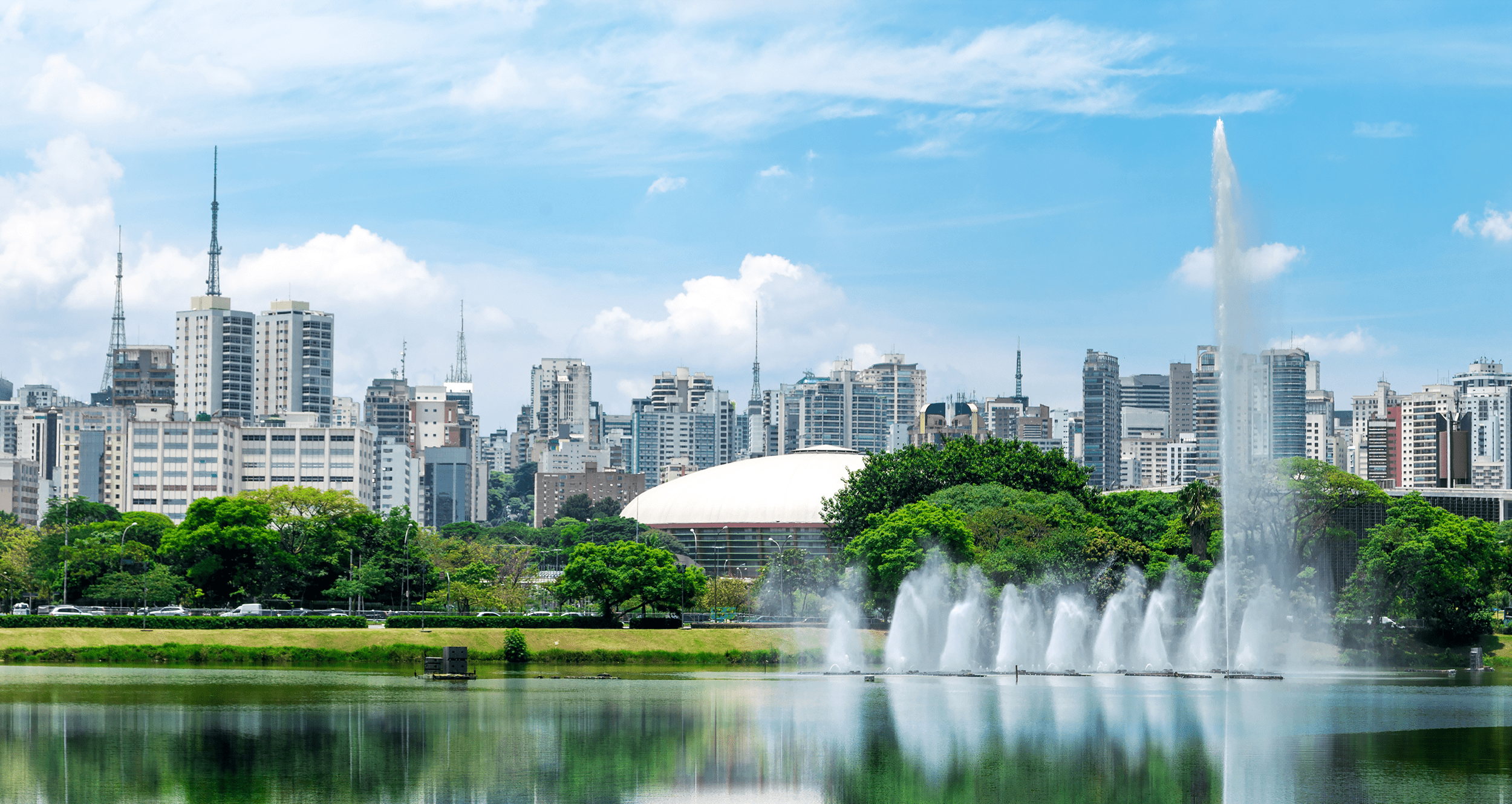 City skyline with tall buildings behind a wide fountain spraying water, and green trees along a lake in the foreground.