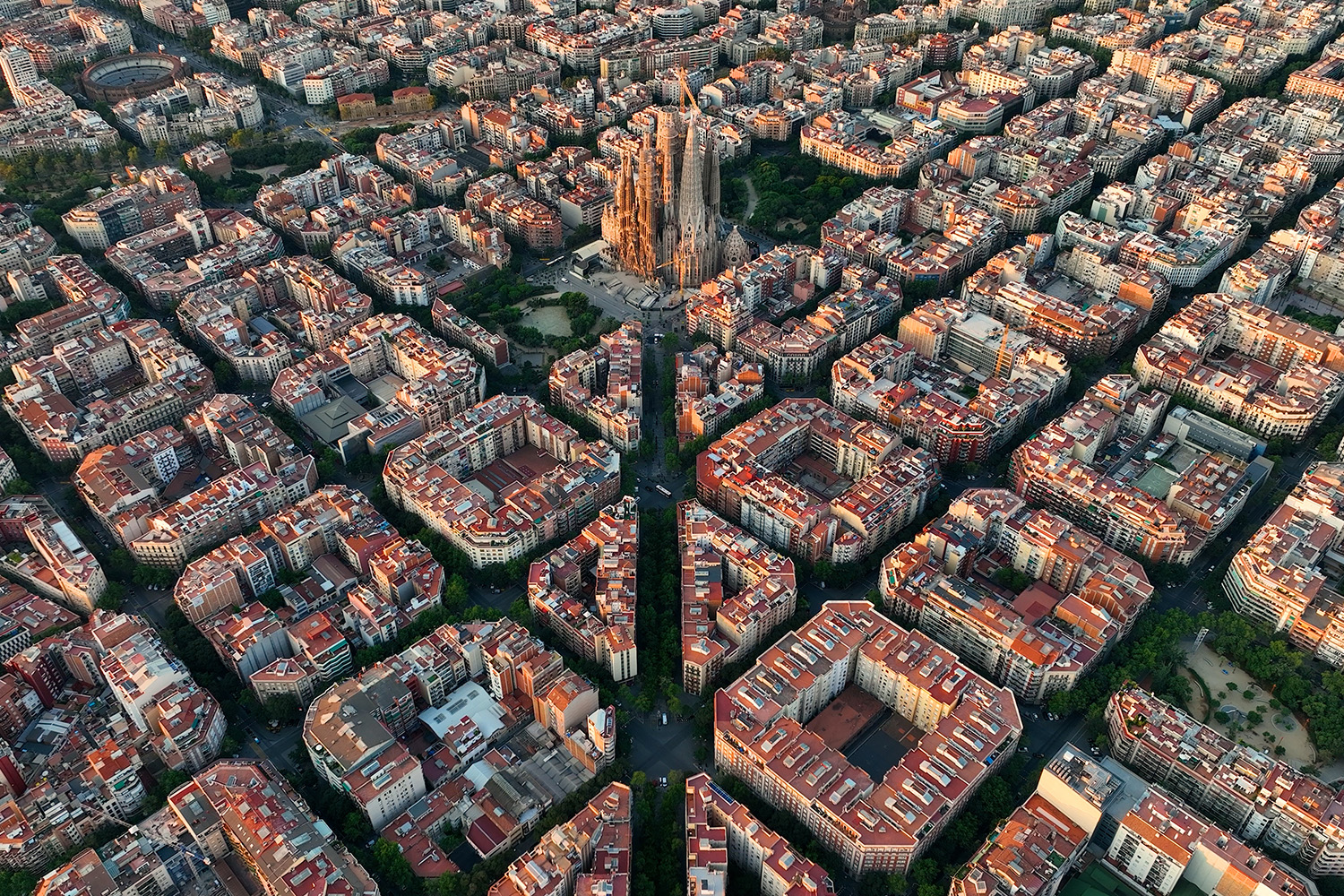 Aerial view of a dense city with a tall, ornate cathedral at the center (Sagrada Família) surrounded by red-roofed buildings in a grid-like layout in Barcelona.