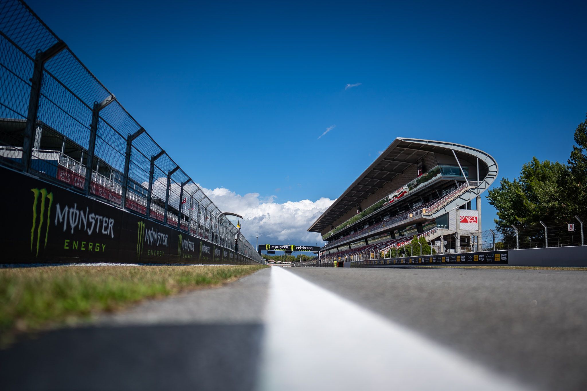 Low-angle view of a race track with safety fences along the left and a curved grandstand on the right under a bright blue sky